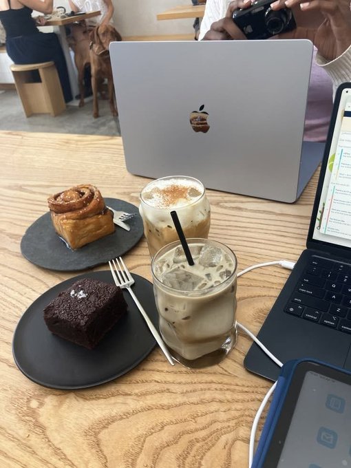 A café scene with a MacBook laptop, an iPad, and two iced coffee drinks alongside a cinnamon roll and a brownie on black plates on a wooden table.