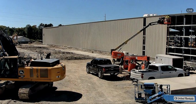 A live construction site showing a large metal building under construction, with heavy equipment including a John Deere excavator, a cherry picker, pickup trucks, and a United Rentals logo visible.