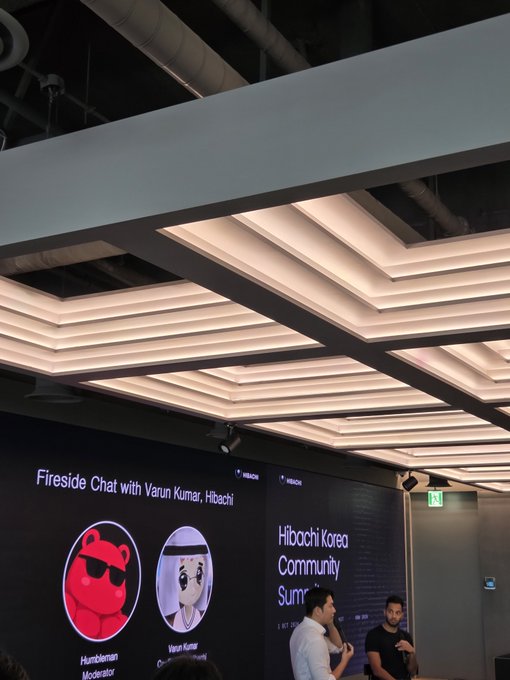 Photo from the 'Hibachi Korea Community Summit' showing a 'Fireside Chat with Varun Kumar, Hibachi' presentation screen with cartoon avatars of a 'Humbleman Moderator' and 'Varun Kumar', while two people speak below.
