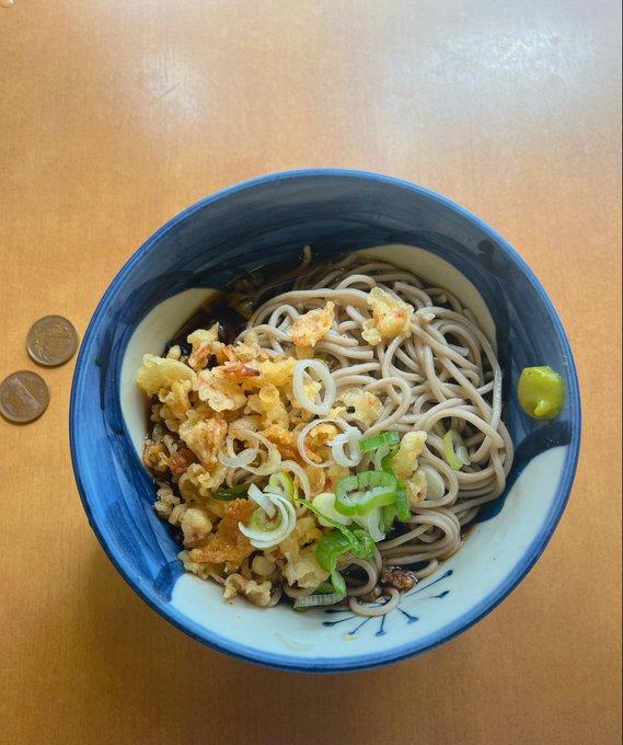 Overhead view of a bowl of soba noodles with tempura flakes, green onions, and wasabi, with two coins beside it for scale.