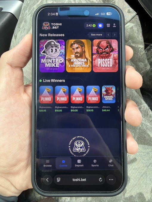 A hand holds a smartphone displaying the Toshi.bet casino app at 2:34, showing a $2.42 balance and featuring new release games like "Minted Mike," "Arizona James & The Lost Relic," and "Pissed," along with live winners on Plinko and Heroic Spins. The sleek UI aligns with the post's claim that Toshi.Bet's design "beats any other casino by miles," supporting the user's excitement about the app update and their hopes for "generational wealth."