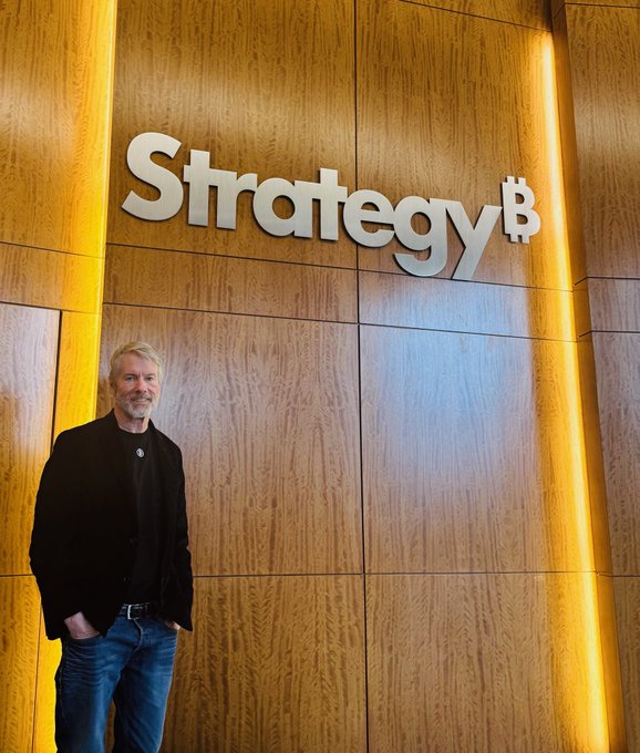 MicroStrategy founder Michael Saylor poses in front of a wooden wall featuring the illuminated 'Strategyᵇ' company logo.