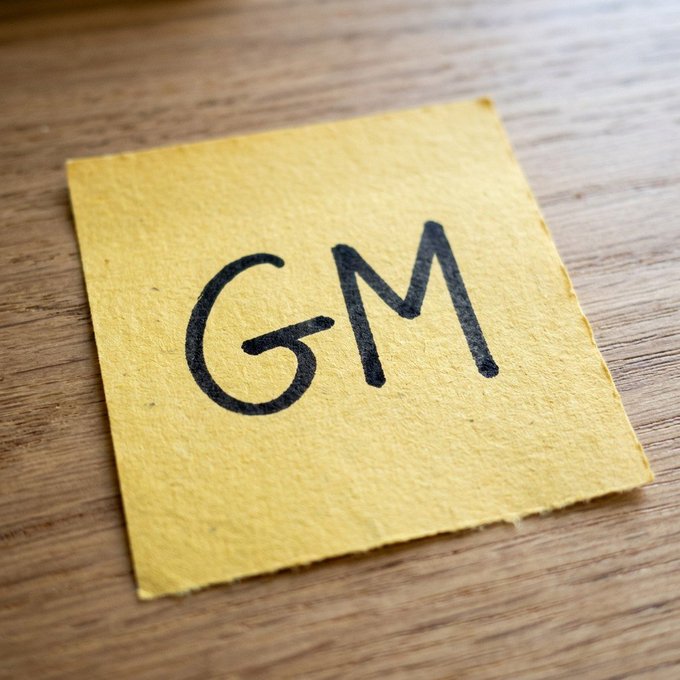 A close-up photo of a yellow sticky note with 'GM' written on it in black marker, resting on a wooden surface.