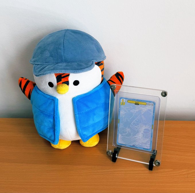 A plush toy dressed in a blue detective hat and vest sits next to a Pokémon card displayed in a clear acrylic case on a wooden surface.