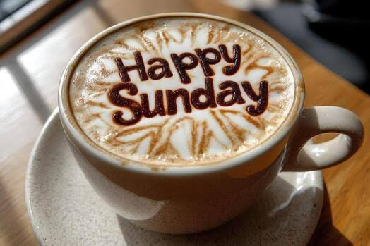 Close-up of a cappuccino in a white cup with chocolate art spelling out 'Happy Sunday' on the foam, served on a saucer.