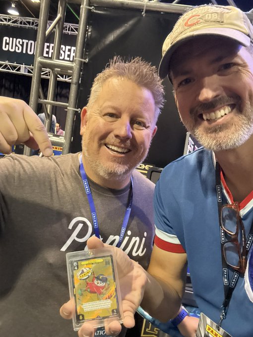 A selfie of two smiling men at what appears to be a convention; one man in a grey 'Panini' shirt holds up a graded trading card in a plastic case, while the other wears a blue shirt, glasses, and a baseball cap.