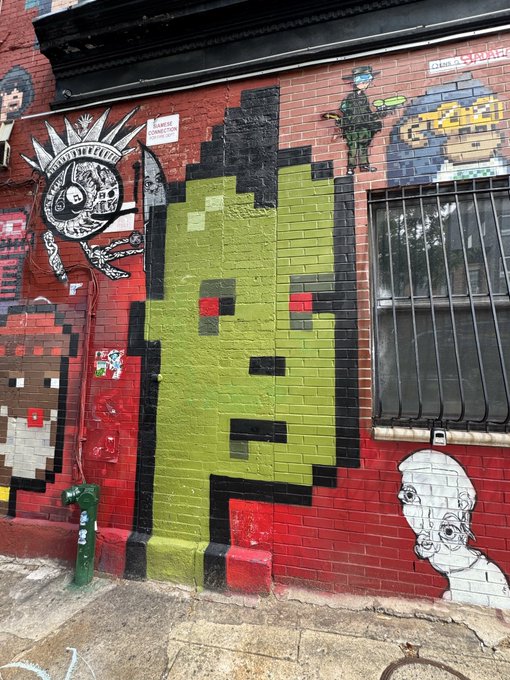 Colorful street art covers a brick wall featuring a large green pixelated face, a tactical figure labeled "CELL," a spiky alien character with "FREE YOUR NERVE ENDINGS" text, and other graffiti, fitting the playful "Ready for summer 26" vibe with its urban, edgy aesthetic.