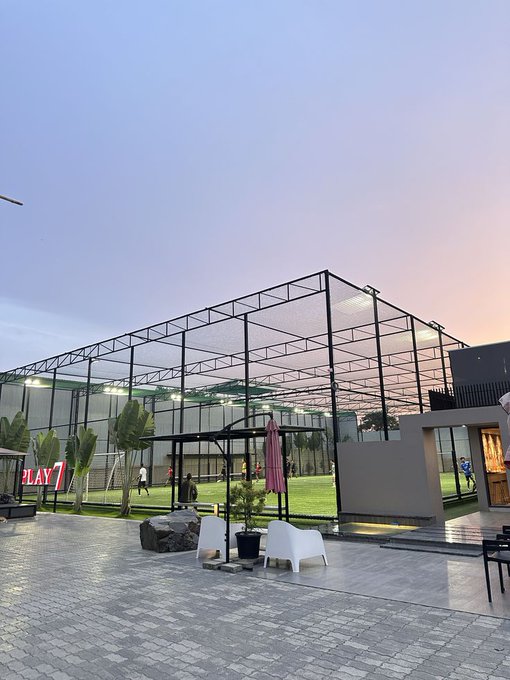 An outdoor sports facility with a high netting enclosure is shown at dusk, with people playing on an artificial grass field and 'PLAY 7' signage visible.