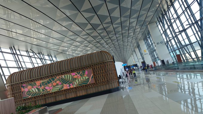 The interior of a modern airport terminal with a geometric patterned ceiling, large windows, and a decorative wooden structure featuring tropical leaf artwork.
