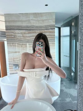 A mirror selfie of a woman in a beige strapless dress inside what appears to be a modern bathroom.