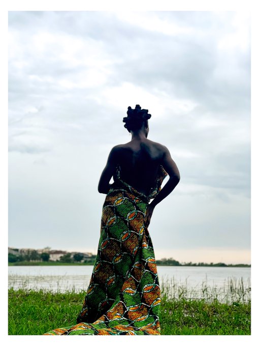 A silhouetted woman wearing a vibrant green, yellow, and orange patterned wrap dress, standing by a lake under a cloudy sky.