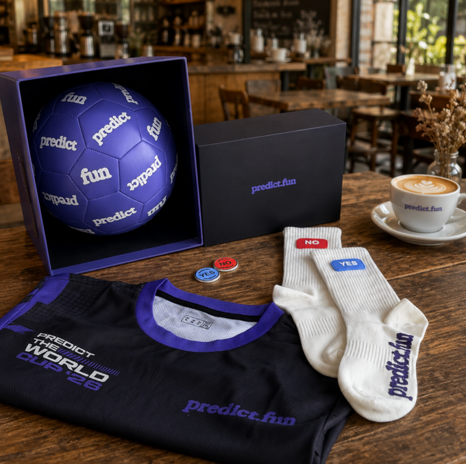A purple soccer ball branded with "predict" and "fun" sits in a black gift box alongside a matching "predict.fun" jersey, white socks, YES/NO pins, and a branded latte, showcasing the World Cup-themed merchandise from @predictdotfun mentioned in the post.