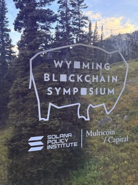The image shows a glass sign for the "Wyoming Blockchain Symposium" featuring the Solana Policy Institute and Multicoin Capital logos, set against a backdrop of evergreen trees and mountains—capturing the scenic setting of the event the author attended.