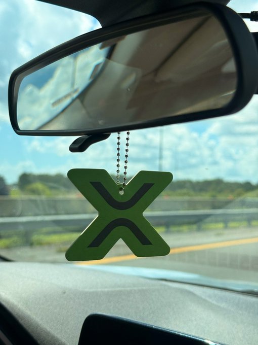 The image features a car's rearview mirror with a green letter "X" hanging from it, possibly as a decorative element or a reminder of something important. The photo also includes text that reads "only a real xrp maxxi owns this freshener". This suggests that the "X" on the mirror is not actually an X but rather a fake one, likely used for decoration or personalization purposes by someone who wants to add a unique touch to their car's interior.

The image also shows a car with its rearview mirror and side view mirrors visible in the background. The scene appears to be set on a road where there is a highway sign present as well.