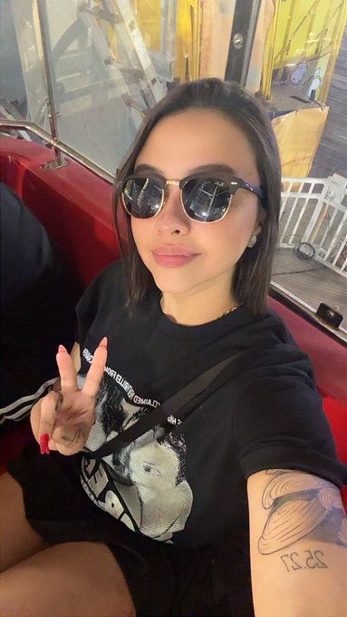 A selfie shows a woman wearing sunglasses and a black t-shirt, with a planet tattoo on her left forearm, sitting near a window.