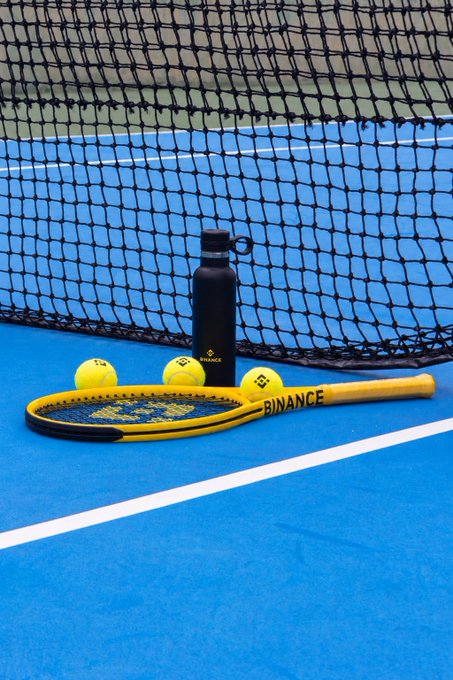 A yellow Binance tennis racket, matching tennis balls, and a black Binance-branded water bottle sit against a net on a blue court, blending crypto branding with sport. The setup mirrors the post's message, pairing Binance's trading hustle with an athlete's winning mentality on the court.