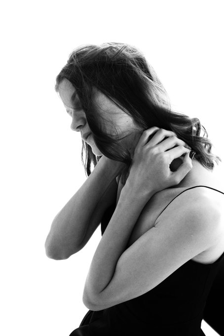 A black and white portrait of a young woman with long dark hair, wearing a black spaghetti strap top, with her arms crossed near her neck as she looks downward.