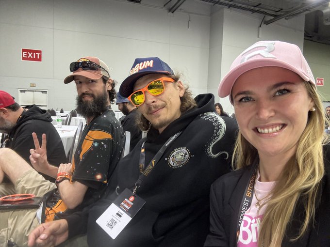 Three people pose for a photo at what appears to be a Bitcoin conference, including a man in a MAGUM cap with orange reflective sunglasses making a peace sign, and a woman in a pink cap wearing a 'MIDWEST BITCOIN' lanyard.