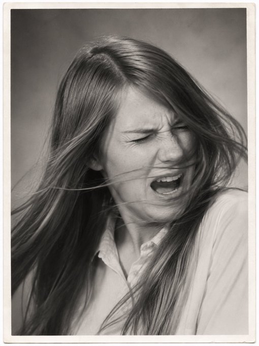 A black and white portrait photograph of a young woman with long hair, eyes shut and mouth open as if yelling or crying, with hair blowing across her face.