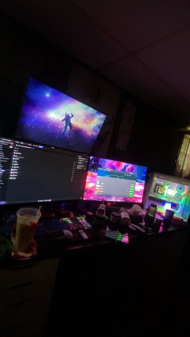 A dimly lit workspace featuring a multi-monitor setup, including a large TV displaying a colorful space-themed wallpaper, a central monitor showing code, and another displaying pink and purple graphics, alongside microphones and a plastic cup.