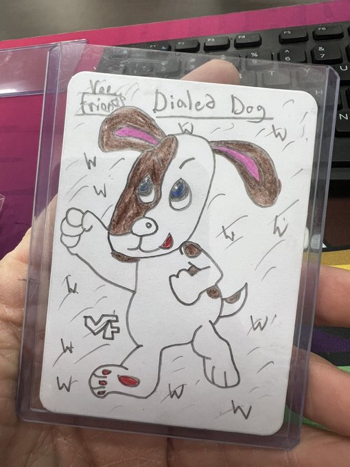 A hand-drawn sketch on a card held in front of a laptop keyboard depicts a cartoon dog with a brown ear, labeled "Dialed Dog" and "Vee Friends"