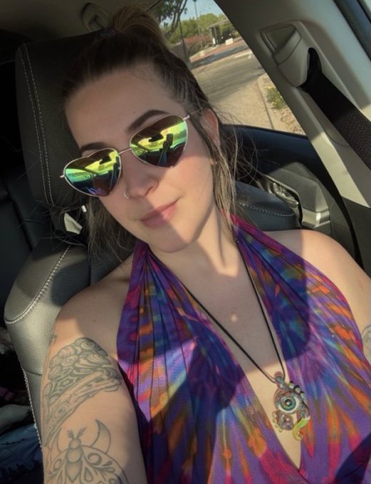 A woman wearing reflective aviator sunglasses and a colorful halter top takes a selfie in a car, displaying a large tree-themed tattoo on her shoulder and a beaded necklace.