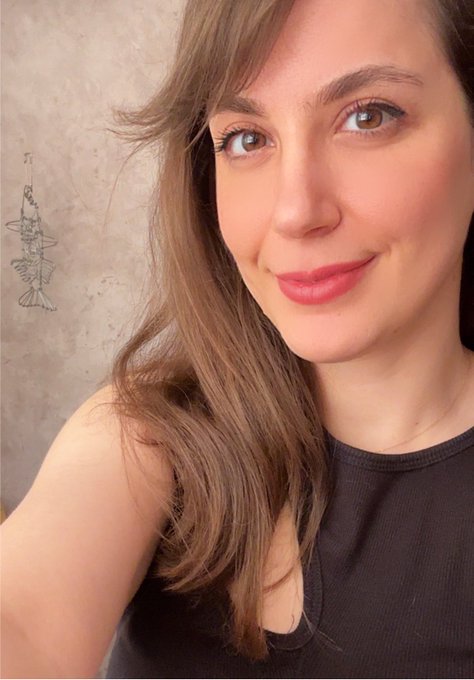 A selfie photograph of a smiling woman with long brown hair wearing a black tank top, photographed indoors against a beige textured wall that features a small decorative hanging ornament.