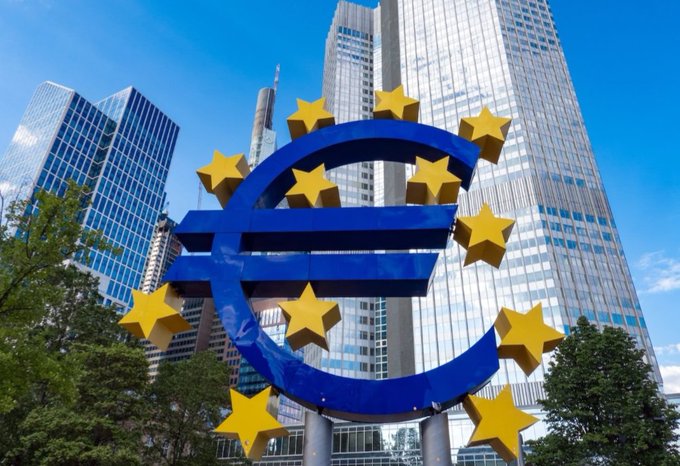 A photograph of the iconic Euro sign sculpture with its circle of yellow stars, located in front of modern glass skyscrapers.