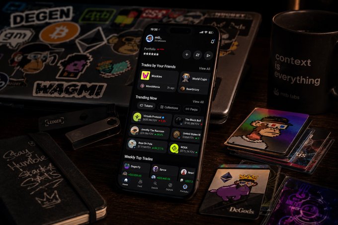 A smartphone displaying a crypto/portfolio trading app sits on a desk surrounded by a "DEGEN" sticker-covered laptop, a Ledger hardware wallet, a mug reading "context is everything web3 labs", and NFT collectible cards.