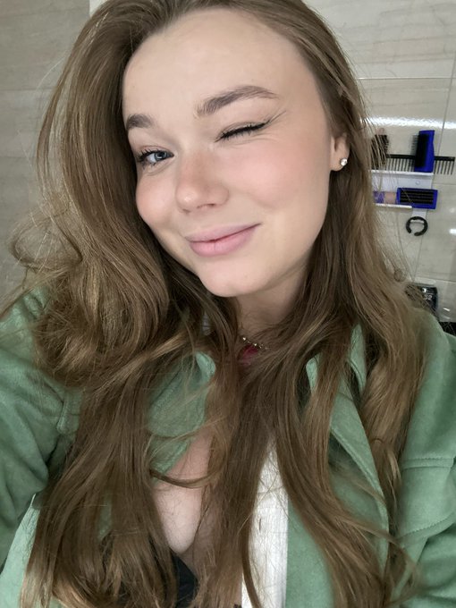 A young woman with long wavy brown hair winks at the camera, wearing a green jacket and a pink top, photographed in what appears to be a bathroom setting.
