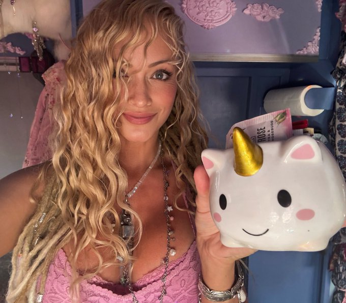 A blonde woman with curly hair holding a white unicorn mug with a gold horn and pink cheeks, standing in a room with a distressed blue door in the background.