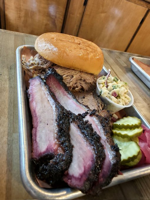 The image features a large tray filled with various types of food, including a sandwich, coleslaw, pickles, and possibly some other dishes. The food appears to be well-cooked and appetizing. The text "Bro, you don’t even know... Who’s hungry? Drop your SOL wallet. I'm buying one person dinner tonight." suggests that the image is related to a social media post or advertisement promoting a meal for someone else.