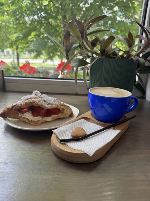 The image features a wooden table with two plates of food - one containing a pastry and the other holding a cup of coffee, both placed near a window. The background shows a potted plant and trees outside the window, creating an inviting atmosphere for enjoying a meal or a break from work. The post includes motivational text encouraging people to have a great week ahead.