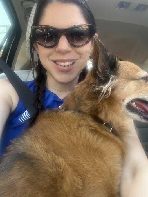 A smiling woman wearing sunglasses and a blue top poses for a selfie in a car with her relaxed golden retriever, enjoying a sunny Sunday morning together.