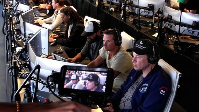 Behind-the-scenes at an esports event with players and commentators sitting at computer setups wearing headsets, with a camera operator filming them in the foreground.