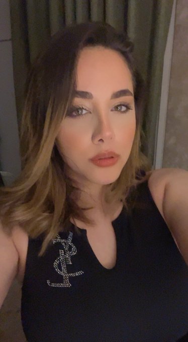 A close-up selfie of a woman with brown hair and dark eye makeup wearing a black top with a rhinestone design.