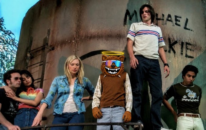 A group of five young people pose in front of a graffiti-covered wall with "MICHAEL" and partial letters "KIE" visible, while a digitally-added cartoon bear wearing sunglasses, a halo, and a brown vest with sharp teeth stands among them—channeling the "fuzzy" vibe and stoner humor of the post's chill, good-vibes message.