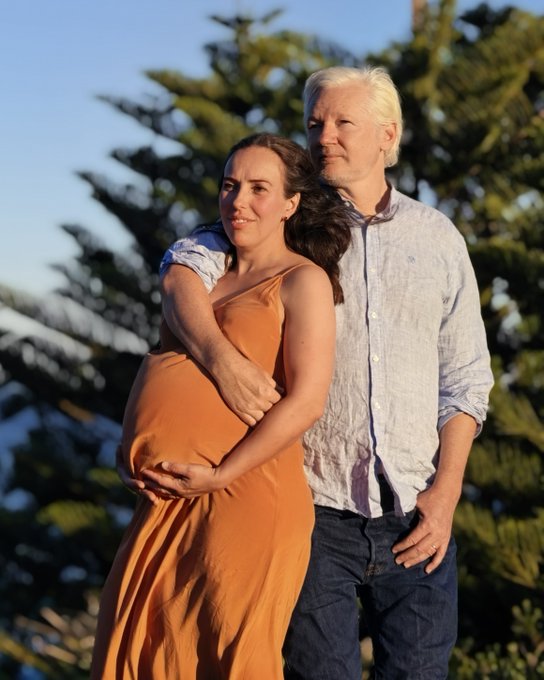 A pregnant woman in an orange dress stands outdoors with a man in a light shirt, who embraces her from behind against a backdrop of pine trees.