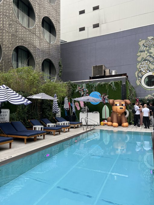 An outdoor pool area features blue lounge chairs, striped umbrellas, and a large inflatable brown bear mascot, with a building featuring circular windows in the background.