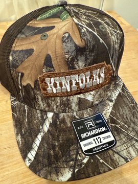 A Realtree-style camouflage trucker hat featuring a leather "KINFOLK" patch is displayed on a wooden surface, with a Richardson 112 cap tag still attached to the brim. This branded merchandise showcases the outdoor aesthetic of the Kinfolks Solana brand.