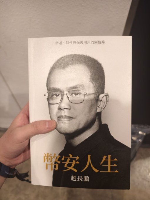 A hand holding up a book titled '酷安人生' by 赵长鹏 (likely a parody of CZ/Binance founder), with text at top reading '幸運、個性與保護用戶的同憶錄'.