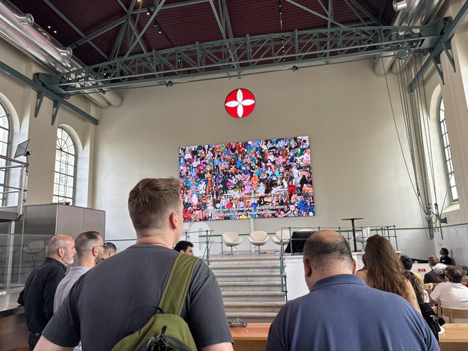 The image shows a group of people gathered around an art exhibit, with some standing closer to the screen while others are further back. The artwork displayed on the wall features a red cross surrounded by a bunch of people. A man wearing a backpack is also present in the scene. The group appears engaged and interested in the art, creating a lively atmosphere.