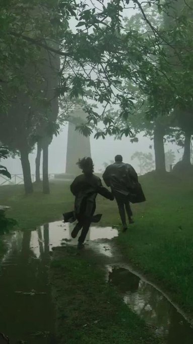 A moody, foggy scene of two people in dark coats running through a wet park path with puddles, surrounded by large trees, with a monument visible in the misty background.