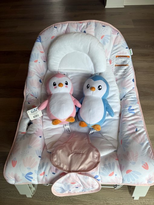 A baby lounger seat featuring two Pudgey Penguins plush toys—one pink and one blue—sitting on a floral-patterned cushion, with a tag reading "Pudgy Penguins" visible on the left penguin, matching the "PenguFather" tweet about the plushie purchase.