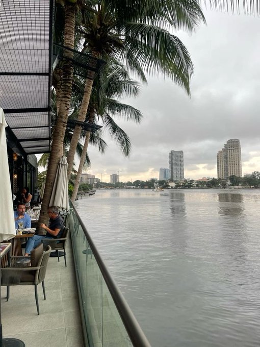 A waterfront café with palm trees and high-rise buildings in the distance, where a few people sit along a railing enjoying the calm river view on an overcast morning. The serene "gm" vibes match the peaceful scene of a slow start to the day by the water.