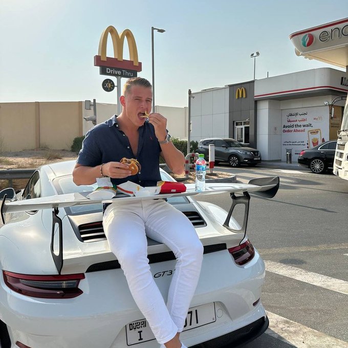 The image shows a man sitting next to his white Porsche sports car, eating a burger while enjoying the outdoors. The scene takes place at a McDonald's restaurant, where there is a sign for Drive-Thru service. The man appears to be in a relaxed and casual setting as he enjoys his meal.