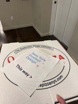 A package sits on the floor containing a white t-shirt and a card for 'The Greenwich Village Institution' with 'New York Product Design' text and a directional arrow pointing 'This way'.