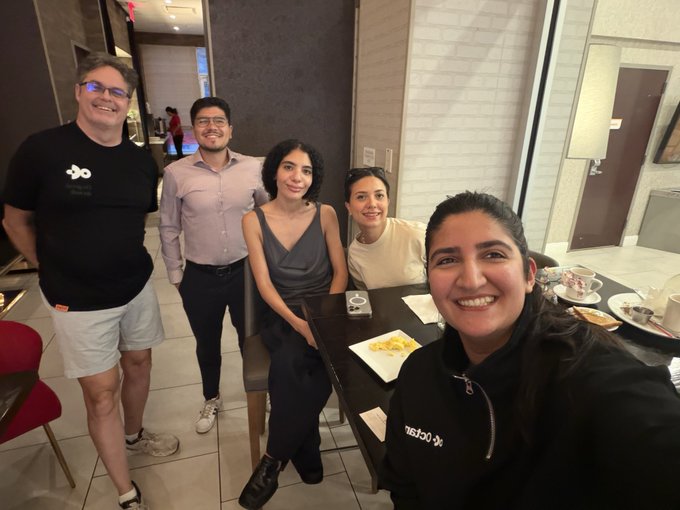 The image shows a group of five people posing for a photo around a dining table with plates of food. The individuals are smiling and appear to be enjoying their time together, possibly at a restaurant or gathering. The background includes a wall clock and a chair, suggesting that they might be in a public space like a restaurant or a social event venue.