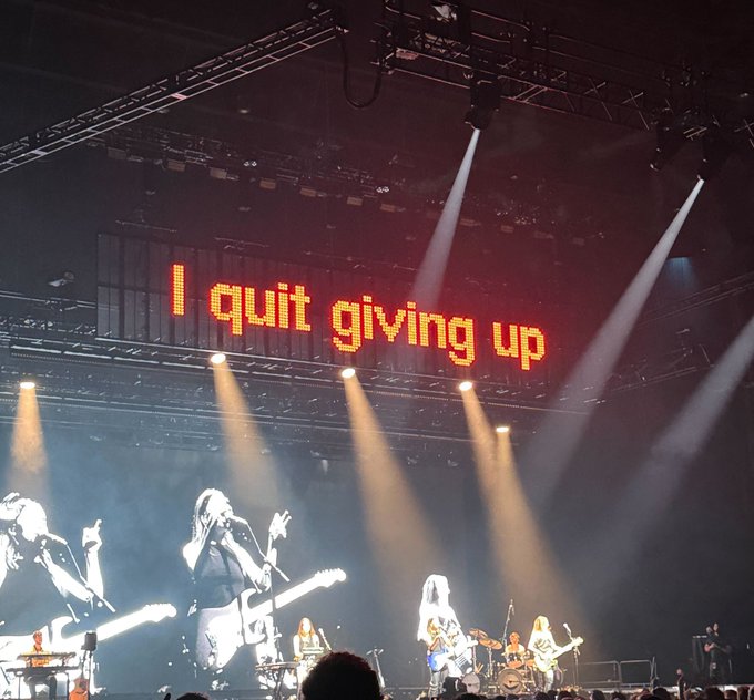 A concert stage with the LED screen displaying 'I quit giving up' in orange text, showing band members performing under dramatic stage lighting.