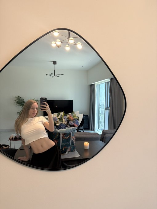 A young woman with long blonde hair takes a mirror selfie while holding a bouquet of flowers, wearing a white top and black skirt in a modern living room with a distinctive curved mirror.